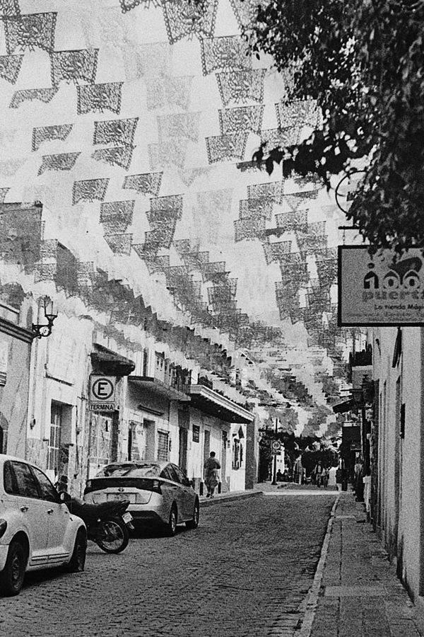 Calle angosta cubierta de papel picado, con autos estacionados